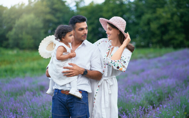 Happy family mother, father and daughter posing in lavender field. Little baby girl wearing straw hat sitting on hands of loving dad, looking at mom outdoors, aromatic flowers. Family, nature concept.